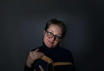 Portrait of a young smiling woman with glasses and a book in hands on a dark background.