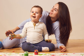 Naklejka premium Happy mother and her little son with paints on his face dressed in home clothes are sitting on the wooden floor in the room and painting with fingers on the white paper