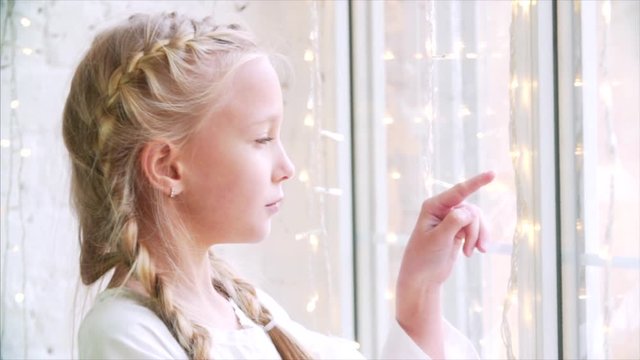 Adorable girl with long braids drawing something with finger on the window. Slow motion. 