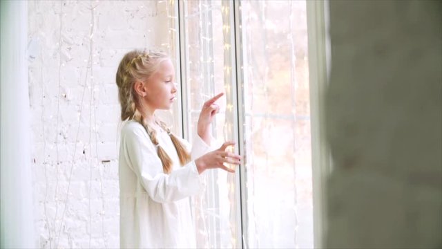 Cute girl with long braids drawing something with finger on the window. Slow motion. 