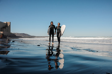 Young couple of surfers walking and laughing along the sea shore with black sand - obrazy, fototapety, plakaty