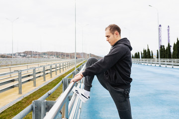 Side view - a handsome slender young guy in a gray tracksuit kneads the joints and ligaments of the legs to a street workout standing on the bridge. Copyspace