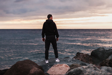 Rear view of fit taut young male in sportswear and sneakers standing on stones by the sea and looking into the sunset on a cloudy summer evening. Concept of sports goal and dream achievement