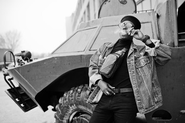 Fototapeta na wymiar African american man in jeans jacket, beret and eyeglasses, smoking cigar and posed against btr military armored vehicle.