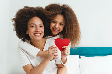 Mother and daughter enjoying on the bed, holding red hearts 