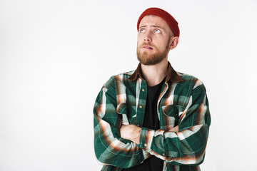 Portrait of young bearded guy wearing hat and plaid shirt looking up, while standing isolated over white background