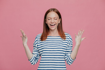 Waist up shot of overjoyed pretty woman gestures with hands, laughs positively, being in high spirit, dressed in sailor striped jumper, closes eyes shut, poses against pink studio wall. Happiness