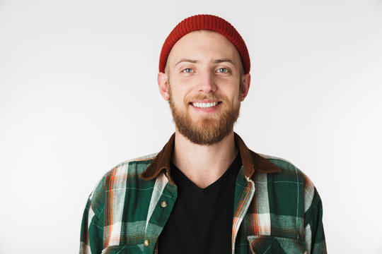 Portrait of stylish bearded guy wearing hat and plaid shirt smiling, while standing isolated over white background