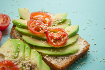 Close up of a sandwich with sliced avocado, tomatoes and sesamum seeds on blue background with copyspace.