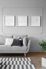 Real photo of a grey living room interior with triptych on a wall, couch and striped rug - obrazy, fototapety, plakaty