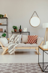 Real photo of bright living room interior with fresh plants, decor on metal racks, lounge with blanket and cushion and mirror on the wall