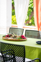 Garden terrace table set up for a lovely brunch on the weekend. Flower petals put in a woven basket to create a romantic, cozy atmosphere. Decorative white and orange curtains behind wicker chairs 