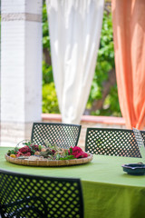 Garden terrace table set up for a lovely brunch on the weekend. Flower petals put in a woven basket to create a romantic, cozy atmosphere. Decorative white and orange curtains behind wicker chairs 