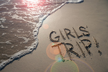 Girls Trip message handwritten on the smooth sand shore of a beach with incoming wave and lens flare