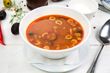 Traditional Russian soup Solyanka cooked with meat, sausages, salted cucumbers and olives. White casserole on the wooden table.