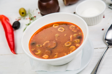 Traditional Russian soup Solyanka cooked with meat, sausages, salted cucumbers and olives. White casserole on the wooden table.