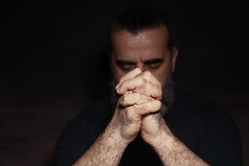 Man bowing his head in prayer with clasped hands in front of his face, in dark room