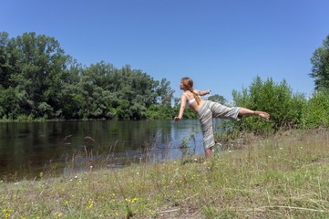 Red-haired girl doing gymnastics on the bank of the river against a background of trees, on a summer sunny day.