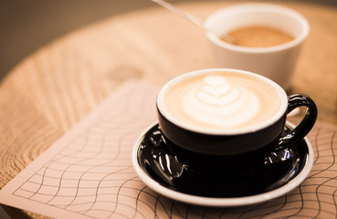 Cup of hot cappucino in a blak cup with coffee art on wooden table background. Hippster style