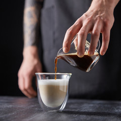 A man's hand pours coffee into a cup of milk on a black wooden table. Cappuccino preparation.
