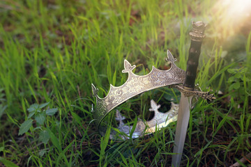 mysterious and magical photo of silver king crown and sword in the England woods or field landscape with light flare. Medieval period concept.
