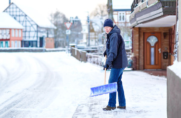 Man with snow shovel cleans sidewalks in winter during snowfall. Winter time in Europe. Young man in warm winter clothes. Snow and weather chaos in Germany. Snowstorm and heavy snowing. Schneechaos