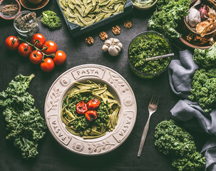 Green pasta with raw kale pesto and grilled tomatoes in plate with fork on kitchen table with ingredients. Vegan food. Healthy meal.  Detox vegetables .  Clean eating and dieting concept. - obrazy, fototapety, plakaty