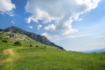 Fototapeta premium Sunny summer day, white clouds over mountain peak, where tourists climb to conquer fear, find courage and develop lateral thinking skills to overcome the difficulty of climbing a mountain. Small road 