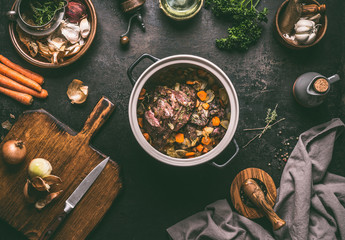 Cooking preparation of stewed meat.  Roasted beef meat in cast iron cooking pot with vegetables on dark rustic background with kitchen utensils and seasoning, top view with copy space.