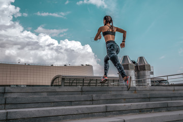 The girl athlete jumps up stairs, runs for a morning jog. In summer in city. The concept of a healthy lifestyle. View from rear on a background of blue sky and concrete stairs. Free space for text.