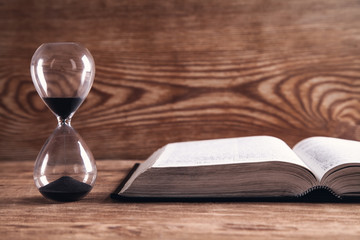 Hourglass with book on wooden table.