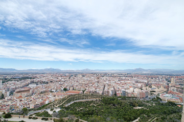 Fototapeta premium Panoramic view of city from Santa Barbara Castle in Alicante, Spain. Block apartment buildings, parks, roads, houses, palm trees. Beautiful mountain landscape in background, blue sky 