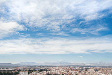 Panoramic view of city from Santa Barbara Castle in Alicante, Spain. Block apartment buildings, parks, roads, houses, palm trees. Beautiful mountain landscape in background, blue sky  