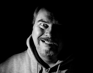 Portrait of bearded adult caucasian man. He smiles like maniac and seems like madness. Black and white shot, low-key lighting. Isolated on black.