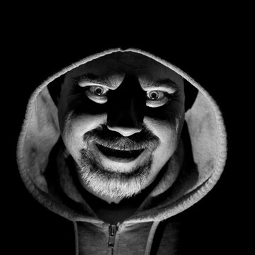 Portrait of bearded adult caucasian man in hood. He smiles like maniac and seems like madness. Black and white shot, low-key lighting. Isolated on black.