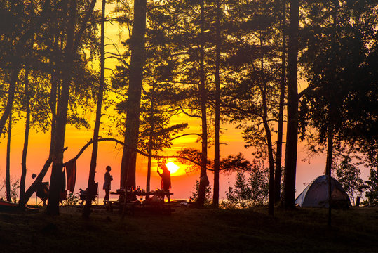 Camping on the beach during the sunset with silhouettes of young people, tents and forest trees. Picnic and barbecue party outdoors. Friends having fun. Concept of relaxation, holidays and recreation