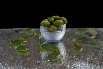 Fruits of green and ripe feijoa in glass bowls with water drops on a mirrored table with green leaves and on a black background