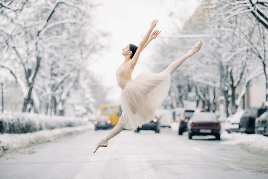 Beautiful ballerina is dancing and jumping on snowy street among cars.