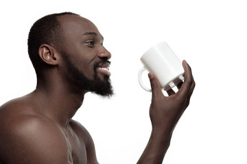 African man with white cup of tea or coffee, isolated on white studio background. Close up portrait in minimalism style of a young naked happy afro man - obrazy, fototapety, plakaty