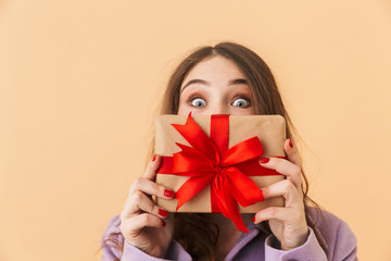 Fototapeta premium Image of surprised woman 20s with long hair smiling and holding present box, standing isolated over beige background