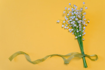 Bouquet of lilies of the valley flowers with green ribbon on bright yellow paper background. Beautiful spring composition in minimal style. Top view, flat lay, copy space