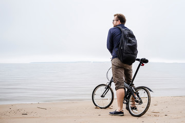 young caucasian man with a small backpack standing with the bike on the beach looking at the sea...