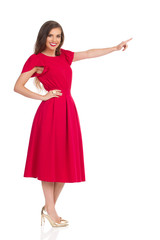 Fototapeta premium Uśmiechnięta młoda kobieta w czerwonej sukience i złotych szpilkach patrzy na aparat i wskazuje