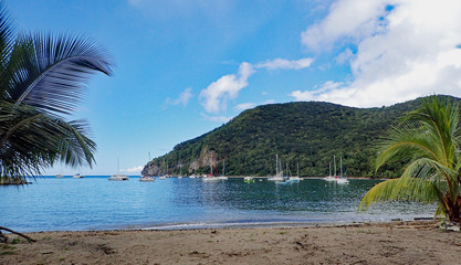 Fototapeta na wymiar Sandy seashore with light surf overlooking an anchorage of sailboats, catamarans, and fishing boats on Caribbean blue waters of a harbor in Daihais, Guadalupe, Windward Islands.