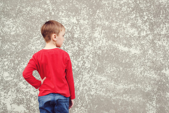 Back view of little boy looking at wall. Rear view on gray concrete wall. Mockup. Cool boy wearing red shirt and jeans. Back to school concept. Copy space.