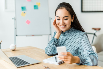 smiling asian businesswoman sitting at computer desk and using smartphone in office