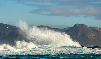 Naklejka premium Big waves break on the rocks in the sea against the backdrop of the coastline. Beautiful seascape. A beautiful moment. Very dynamic photo. Cape Town. False Bay. South Africa.