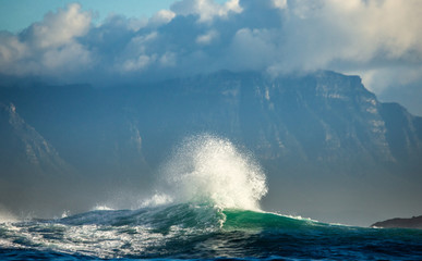 Big waves break on the rocks in the sea against the backdrop of the coastline. Beautiful seascape.  A beautiful moment. Very dynamic photo. Cape Town. False Bay. South Africa.