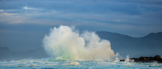 Big waves break on the rocks in the sea against the backdrop of the coastline. Beautiful seascape.  A beautiful moment. Very dynamic photo. Cape Town. False Bay. South Africa.