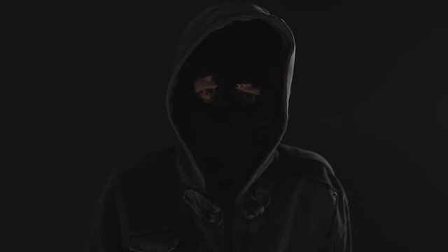 Burglar intruder with flashlight in dark, spooky hooded masked person searching the room with torch and approaching the camera